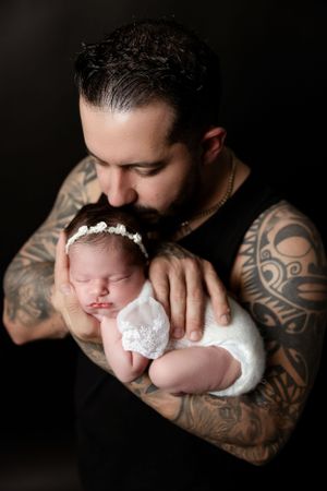 A sleeping newborn in white wrap is held by tattooed arms against dark backdrop.