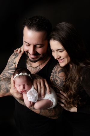Parents with tattoos share joyful smiles while cradling their newborn baby in a dark studio setting.