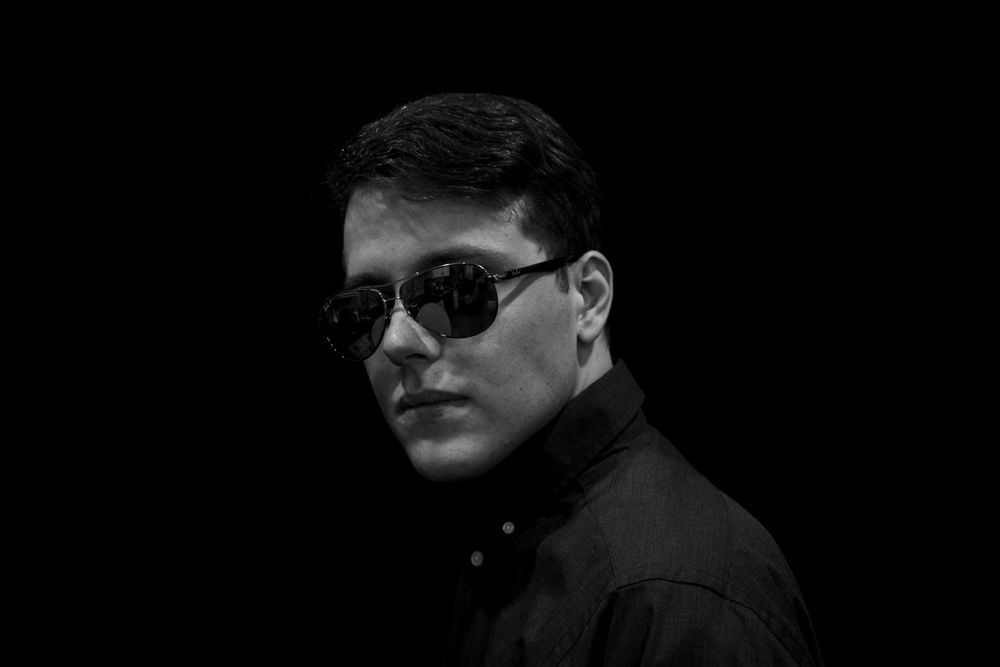 Dramatic black and white portrait of person wearing sunglasses and dark clothing against dark backdrop.