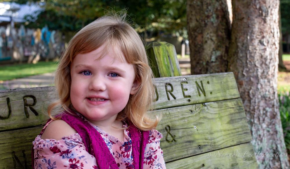 Young child smiling in front of a wooden sign outdoors.