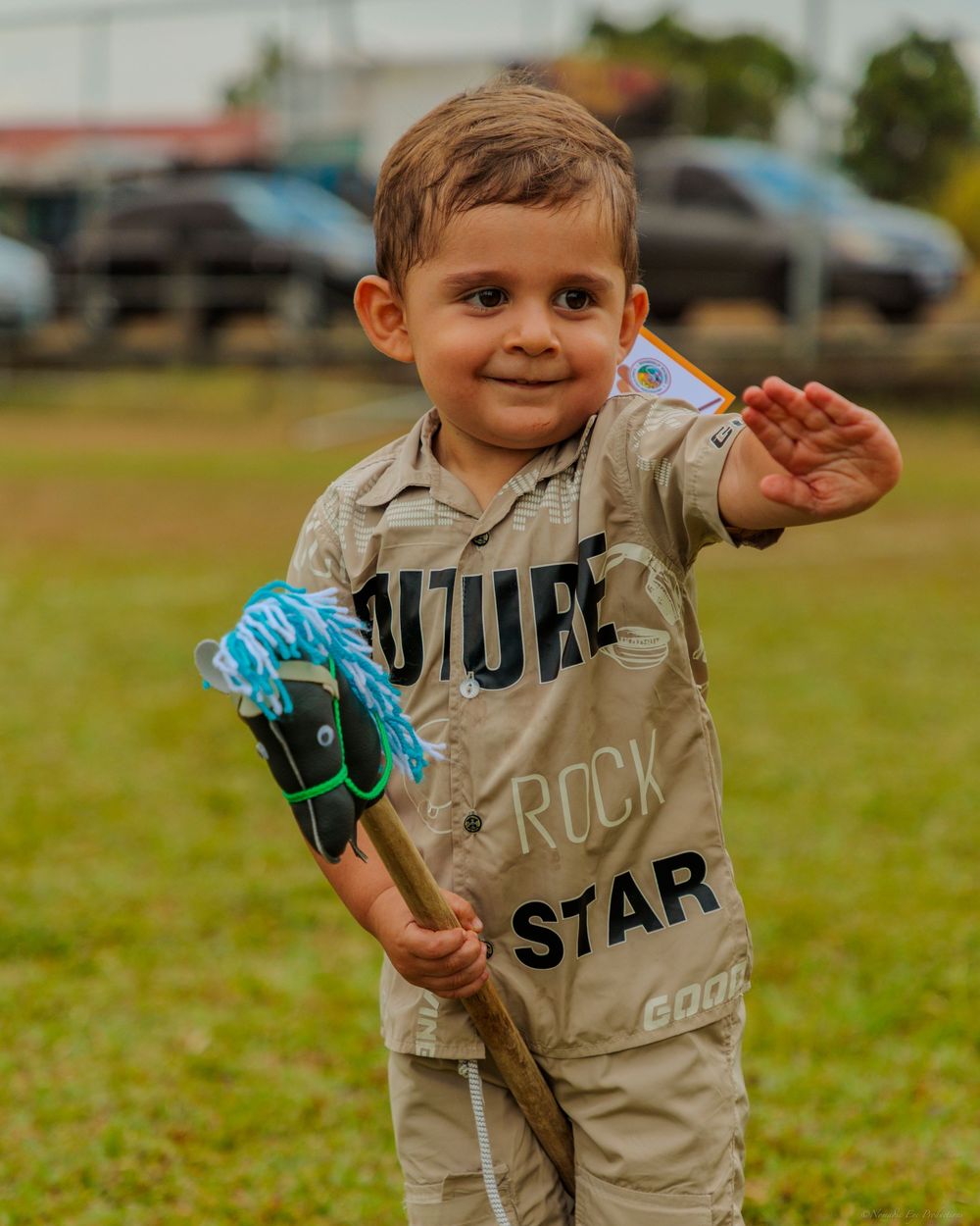 A smiling child in a beige uniform waves while holding a blue lacrosse stick on a grassy field.