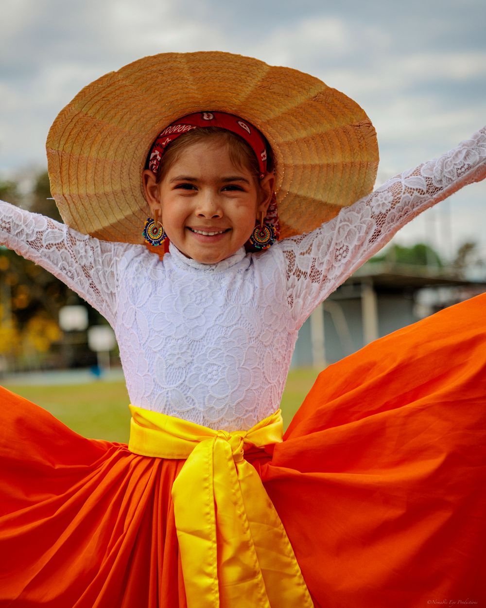 Traditional folk dancer poses outdoors wearing bright orange skirt, white lace top and wide straw hat.