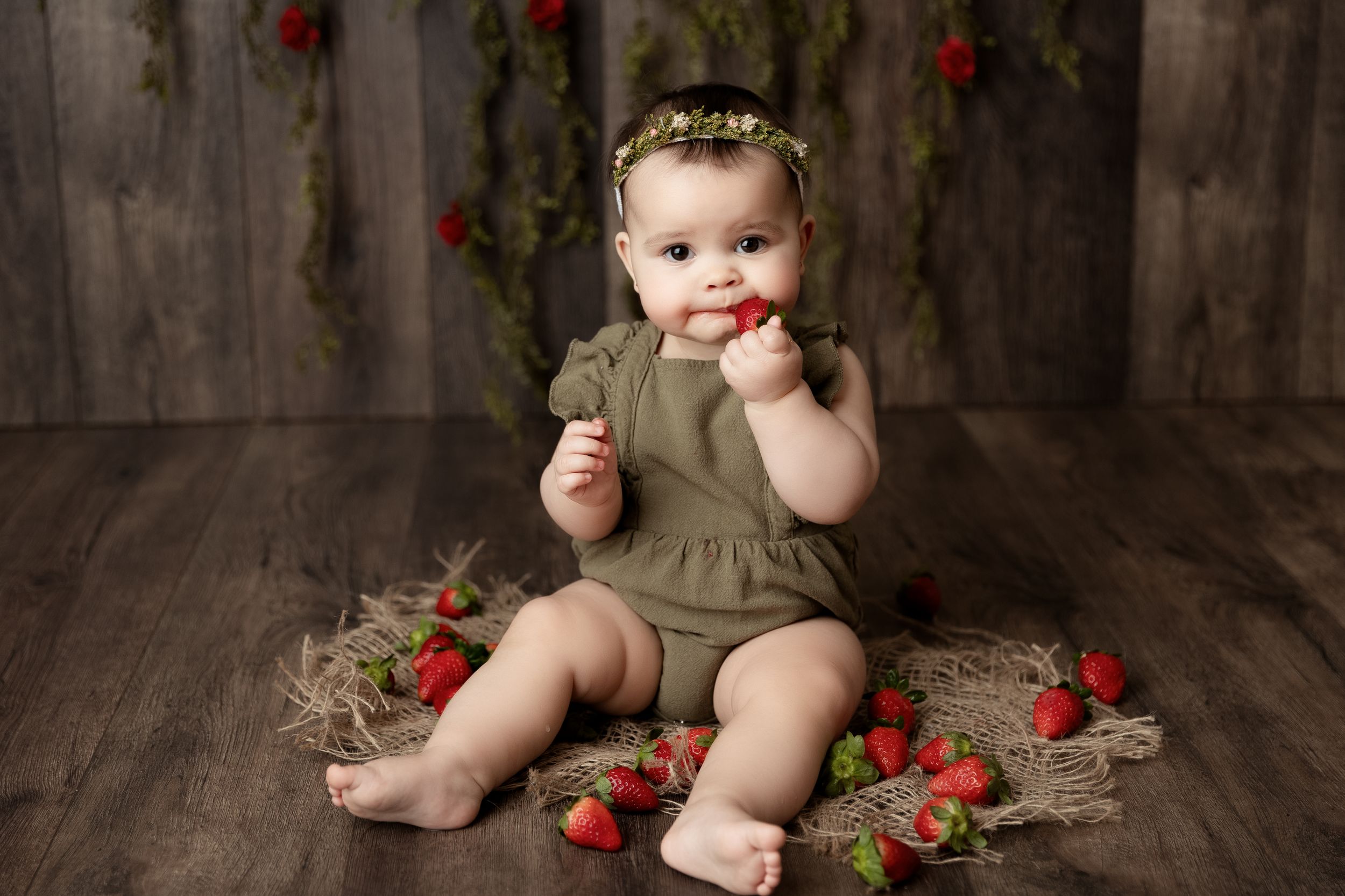 Baby in olive green dress sits on wooden floor surrounded by fresh strawberries during rustic photo session.
