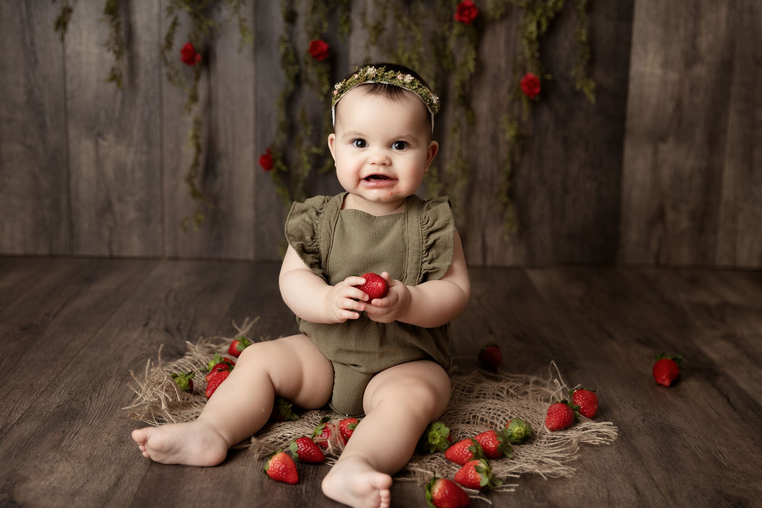 Baby in olive romper sits on wooden floor surrounded by scattered red strawberries in rustic studio setting.