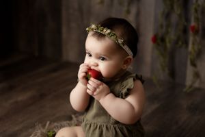 Baby wearing an olive green dress and floral crown sitting against a dark background.