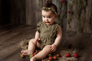 Baby in olive green ruffled dress poses with strawberries on dark wooden floor against rustic backdrop.