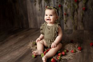 Baby in olive green romper sits on wooden floor with scattered red rose petals in rustic studio setting.