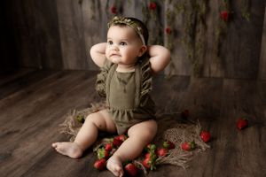 Sweet baby in olive romper sitting on wooden floor surrounded by scattered red flowers in rustic setting.