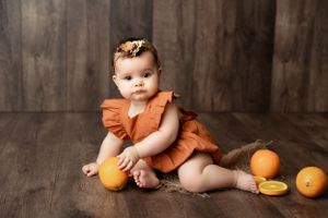 A small child in a rust-colored outfit sits on wooden floor with oranges around them in a rustic studio setting.