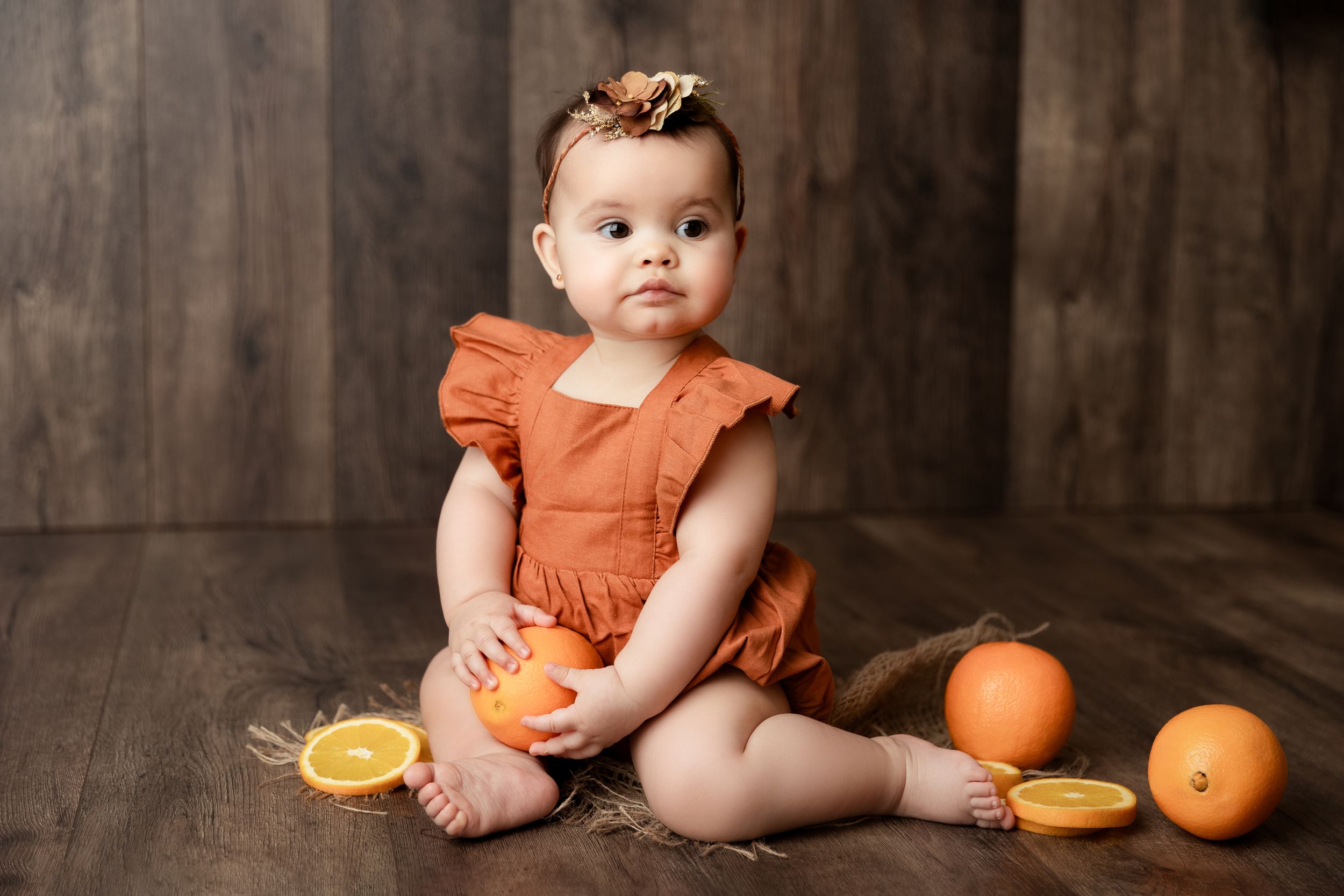 Baby in orange dress sits on wooden floor with oranges and citrus fruit in rustic studio setting.