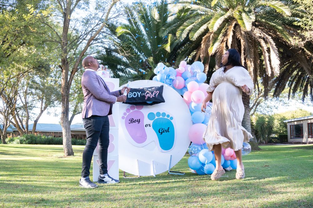 Outdoor gender reveal party setup with balloon arch and footprint banner against palm trees.
