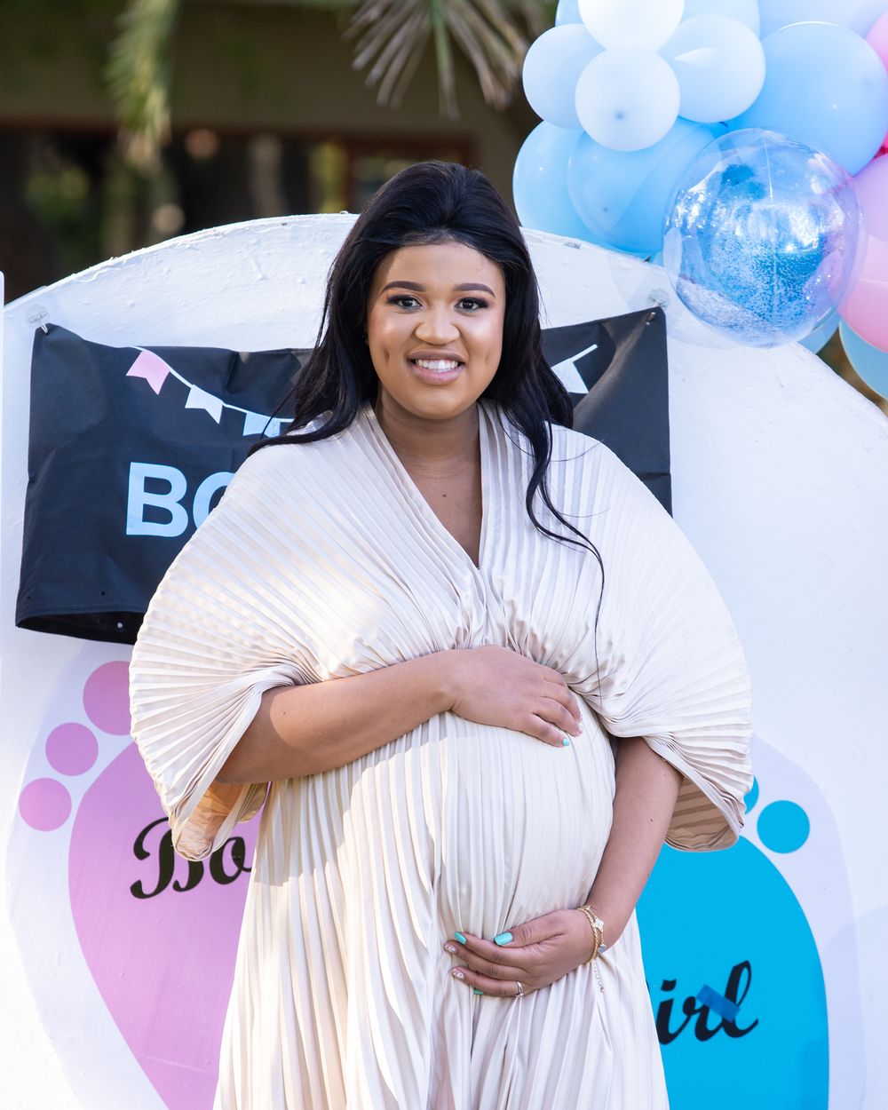 Expectant mother in white pleated dress poses by gender reveal party decorations.