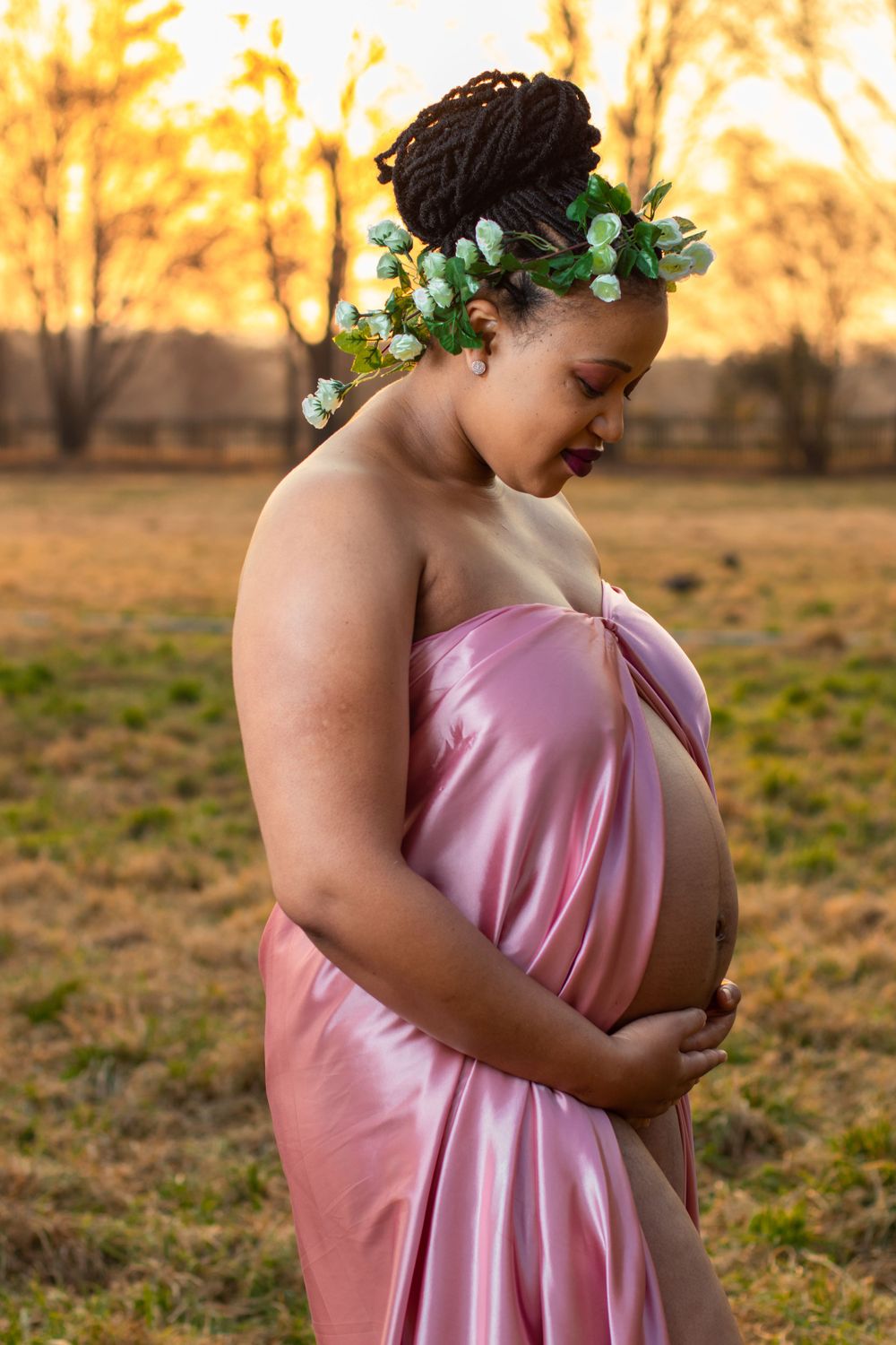 A maternity photoshoot at sunset featuring a pink silk dress and floral crown in a natural outdoor setting.