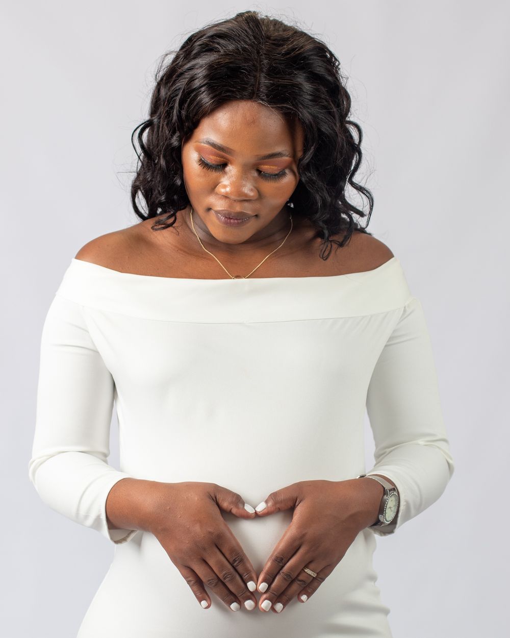 Serene maternity portrait showing mother-to-be in white off-shoulder dress making heart shape over belly.
