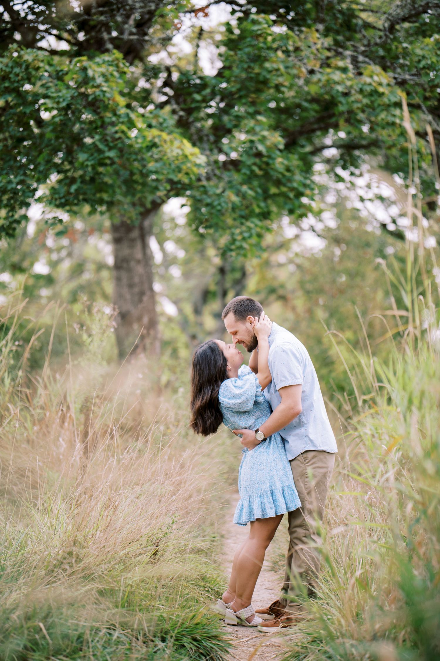Romantic couple embraces in tall grass under a tree in a dreamy outdoor setting.