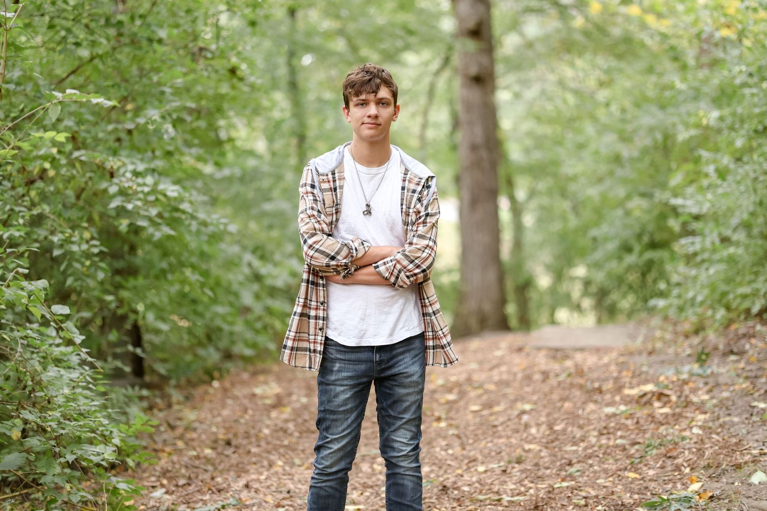Young person in plaid shirt and white t-shirt standing on wooded path.