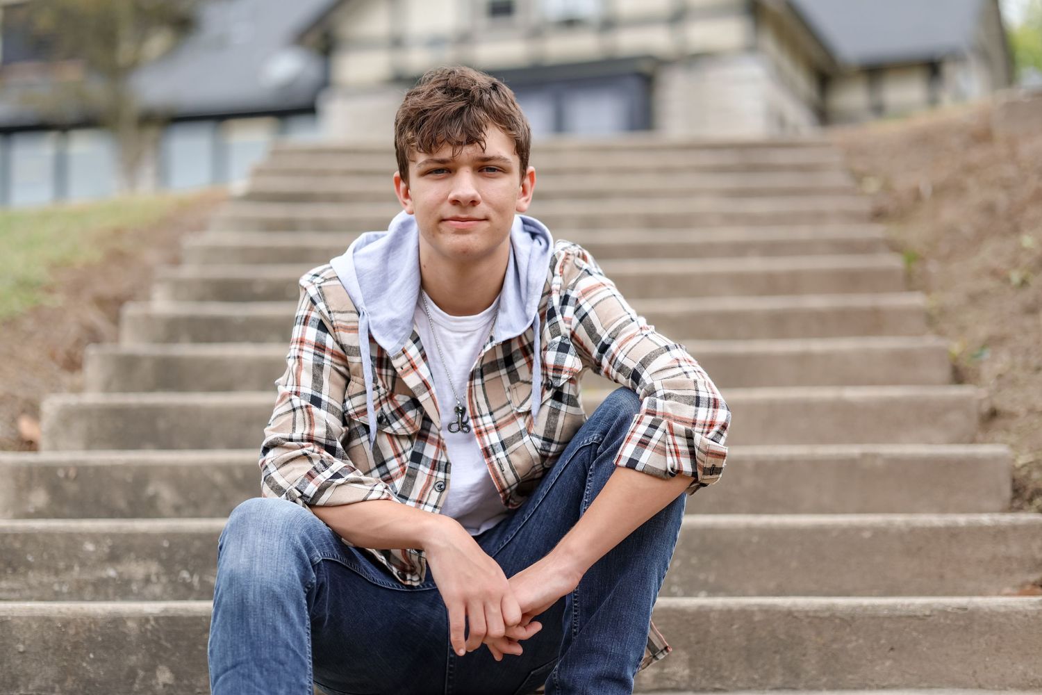 A person in a plaid shirt and jeans sits casually on concrete steps outdoors.
