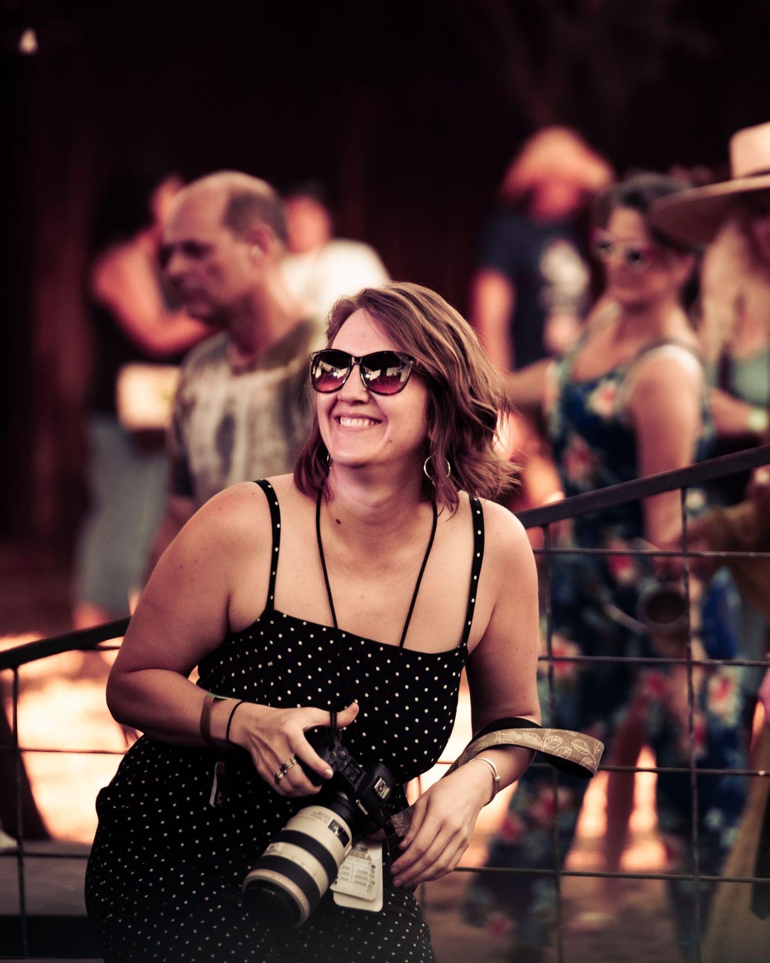 A photographer with a professional camera smiles while wearing a polka dot dress at an outdoor event.