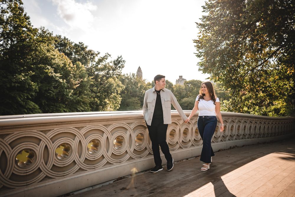 A couple walks along an ornate stone bridge railing on a sunny autumn day in a park setting.
