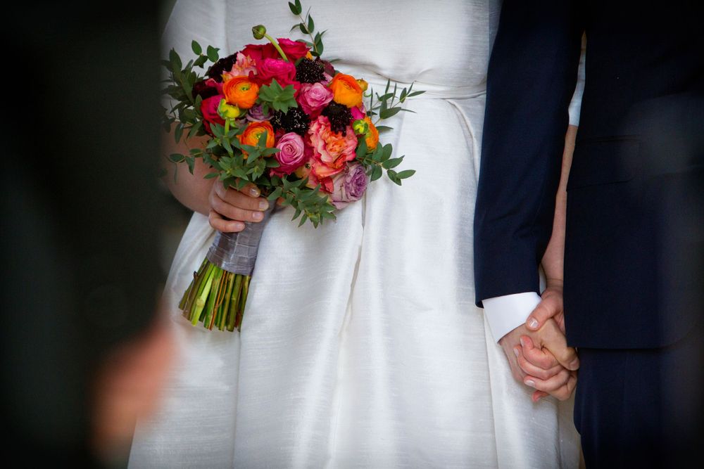A vibrant wedding bouquet of red, orange and purple flowers held against a white dress.
