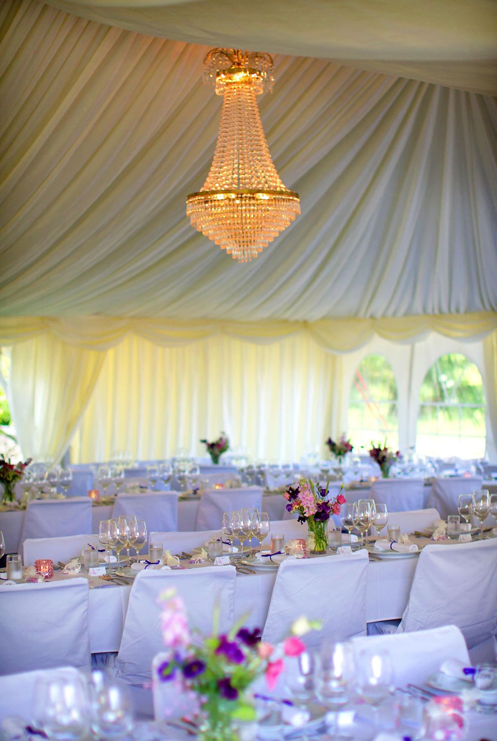 Elegant wedding reception tent with crystal chandelier and purple floral centerpieces.