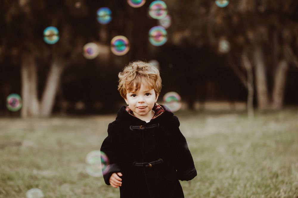Soap bubbles float through the air as a small child in black clothing plays outdoors.