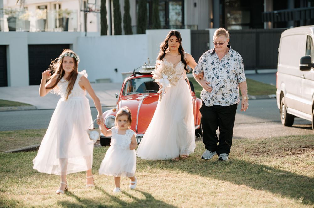 Wedding party walks past red vintage car on sunny outdoor path.