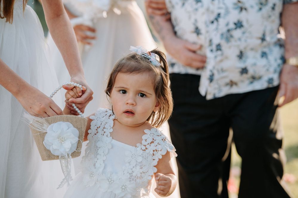 Young flower girl in white lace dress at outdoor wedding ceremony.