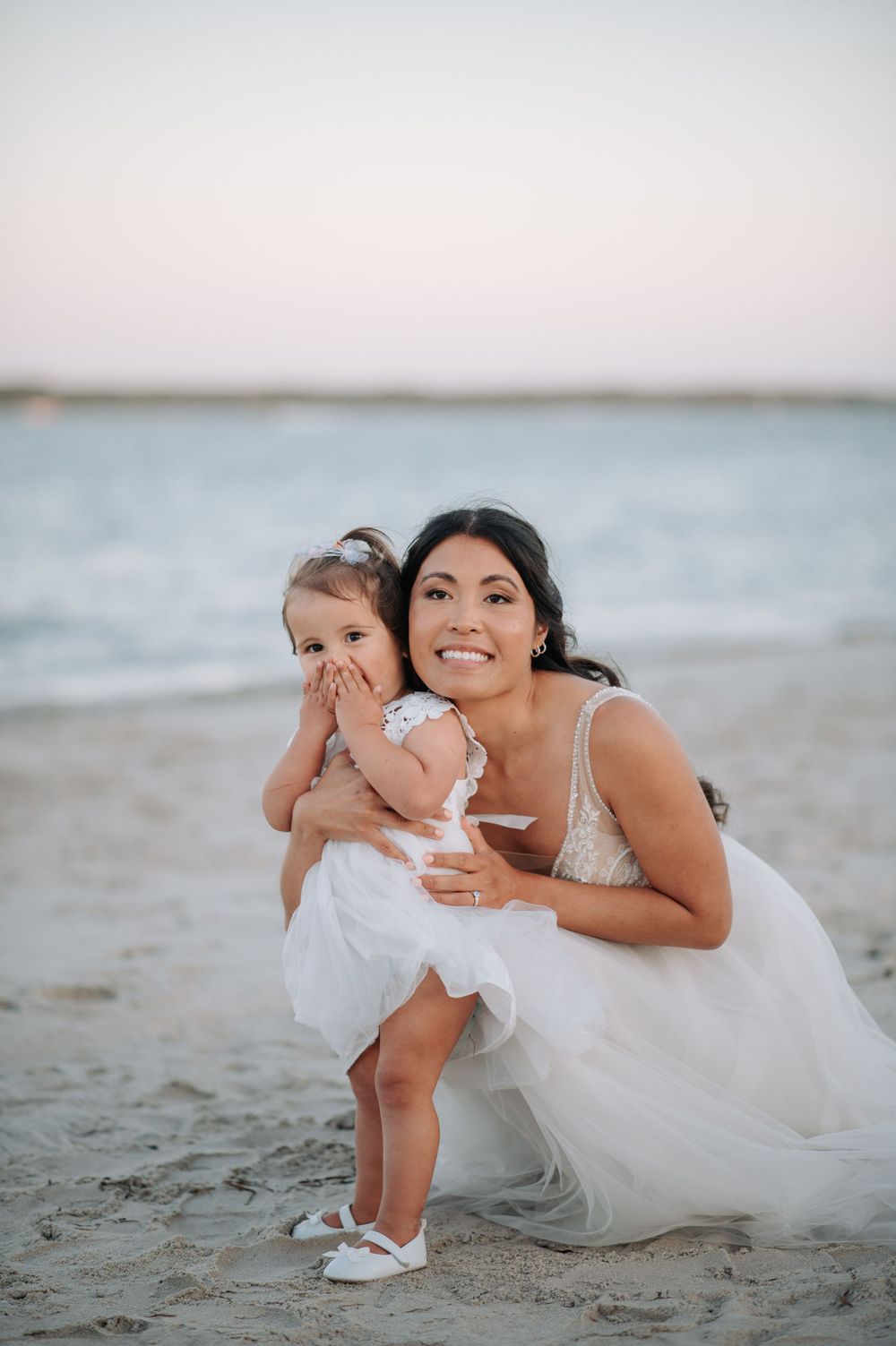 A touching beach portrait of a bride and small child in white dresses against an ocean backdrop.