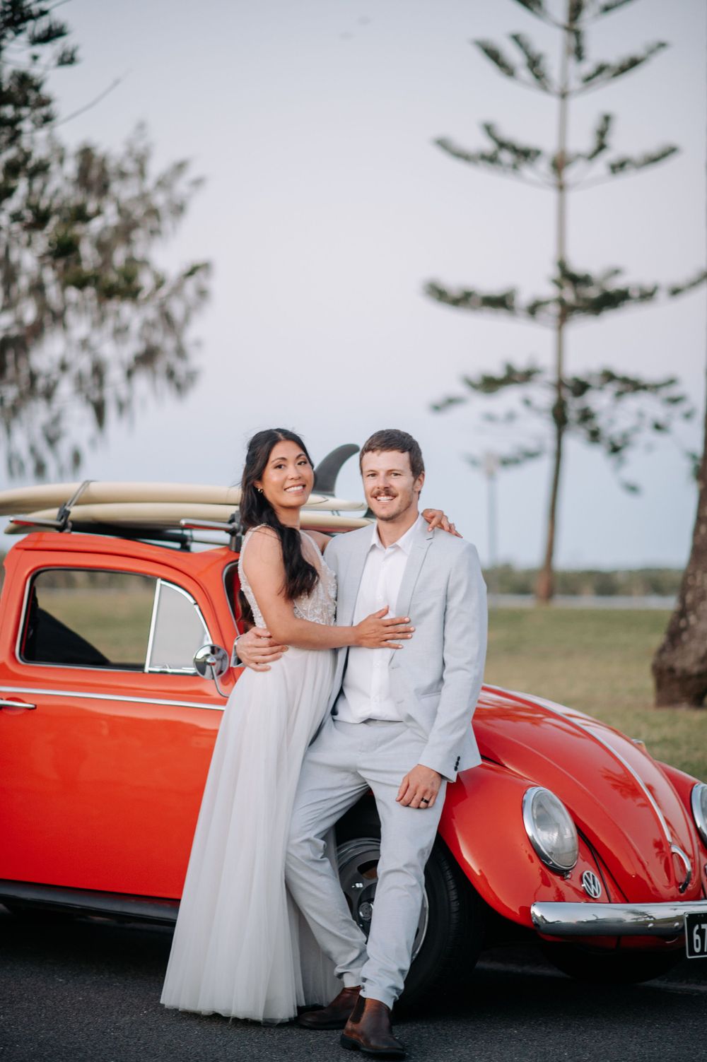 A newlywed couple posing with a vintage red Volkswagen Beetle with surfboards mounted on top.