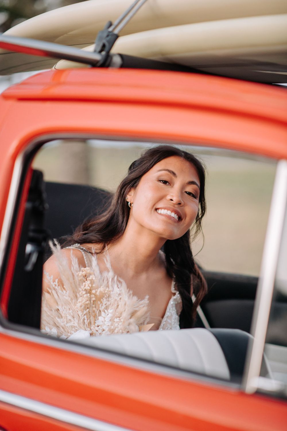 A bride in lace dress smiles from inside a classic red vintage car.