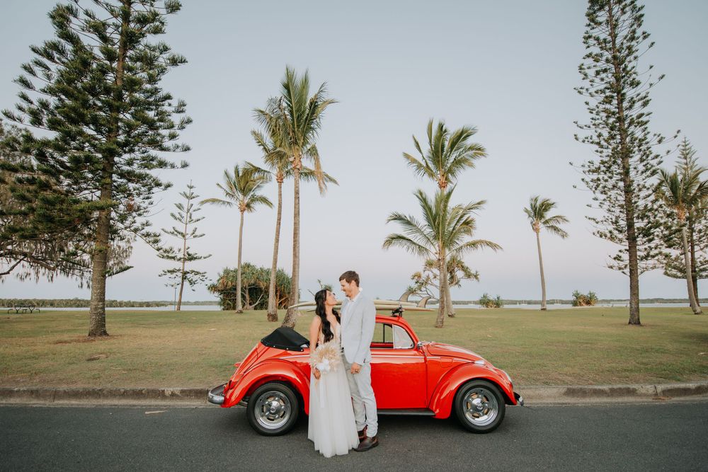 A classic red Volkswagen Beetle parked under palm trees beside a beach pathway during a sunset wedding photoshoot.