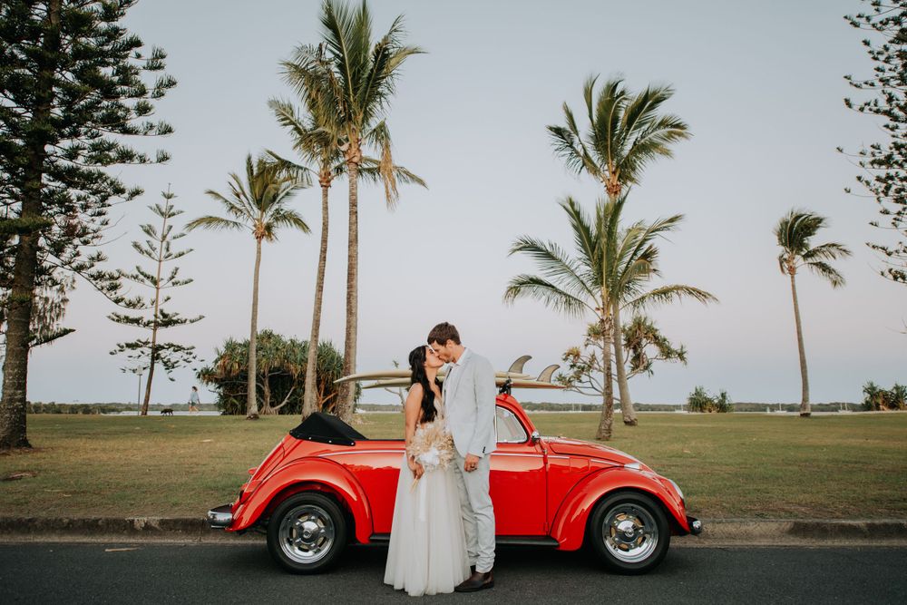 A newlywed couple pose with a vintage red Volkswagen Beetle against a tropical backdrop.