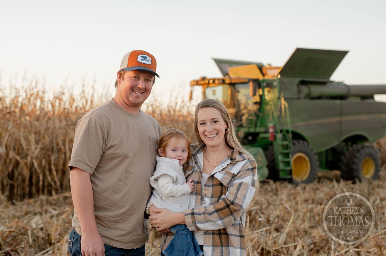 Family stands together near John Deere combine with cornfield in background at sunset.