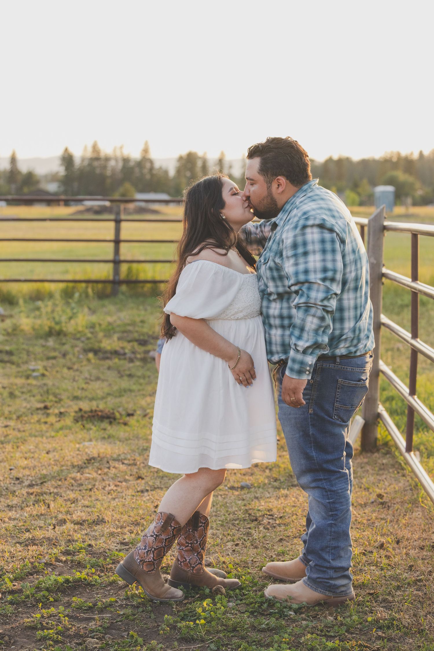 Couple kissing near a ranch fence at golden hour, woman in white dress and cowboy boots.