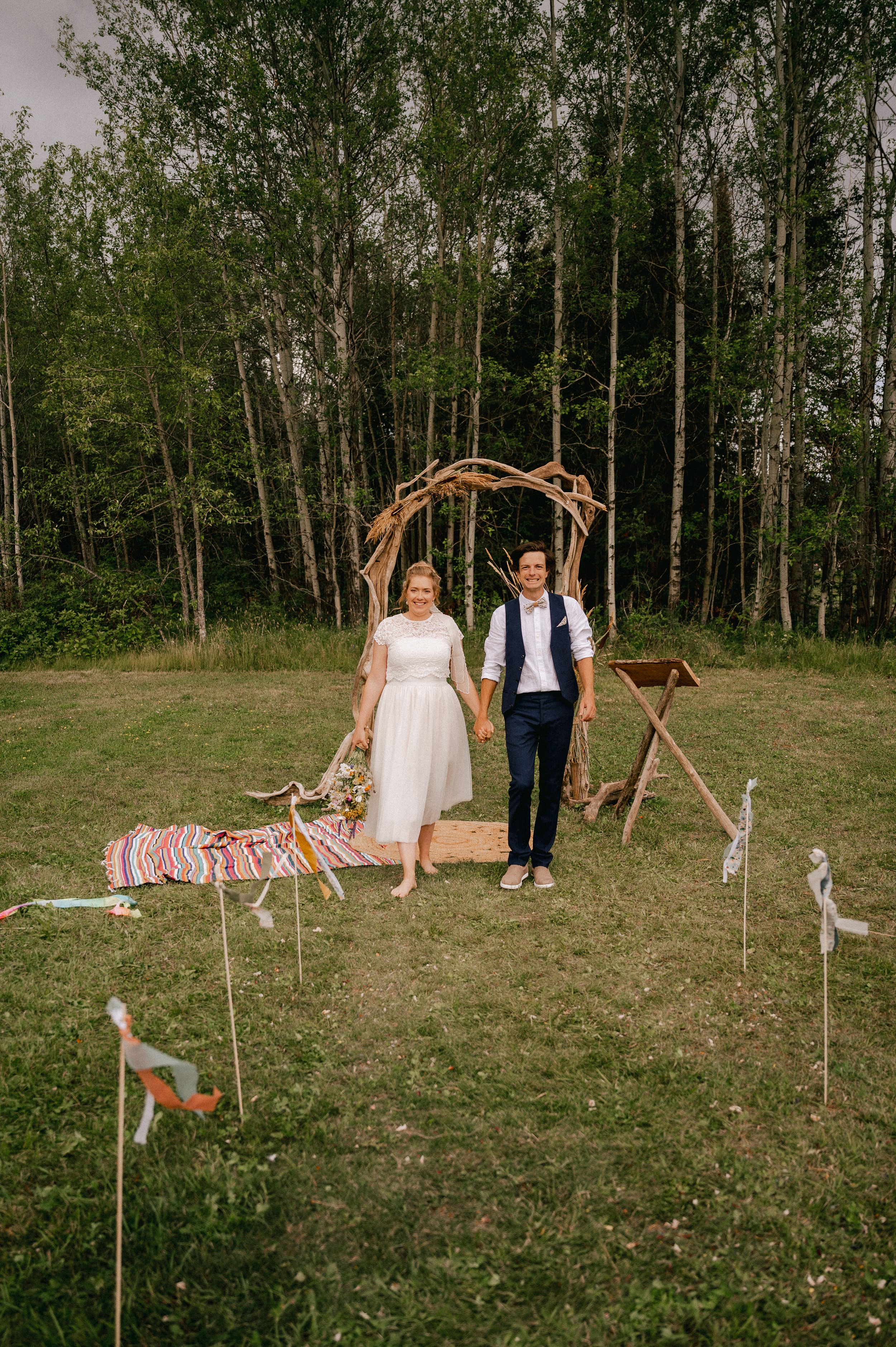 A couple stands in a grassy field near birch trees with a rustic wooden swing set in their outdoor wedding venue.