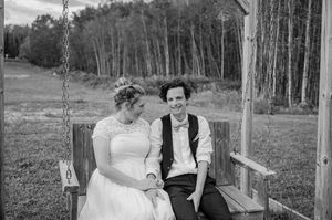 Two people sit together on a wooden swing in front of a forest of birch trees in black and white photograph.