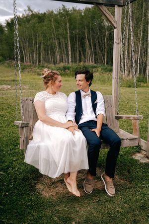 A couple sits together on a wooden swing outdoors with birch trees in the background.