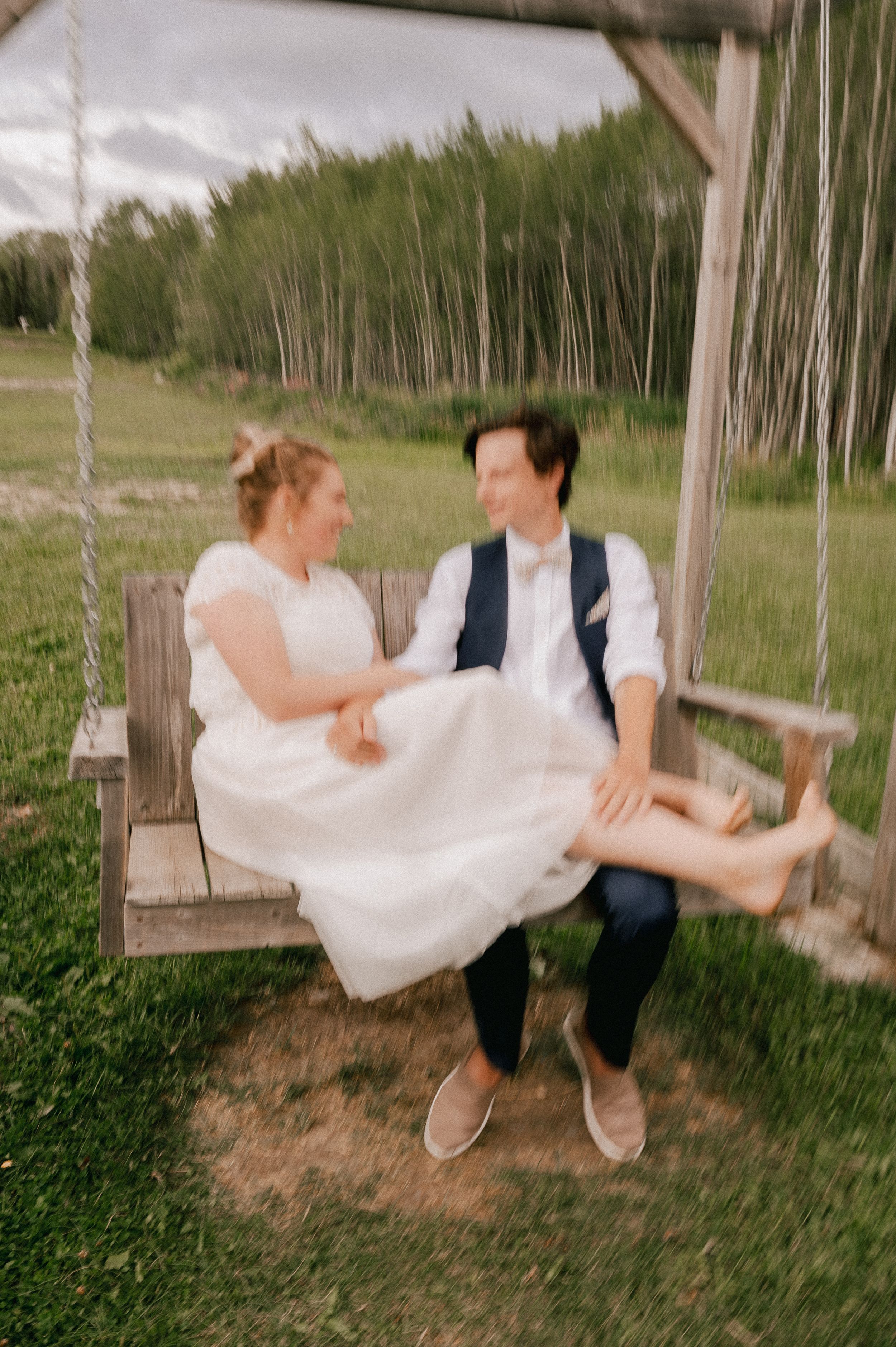 A couple playfully swings together at their outdoor wedding, with the bride's white dress flowing in the motion.
