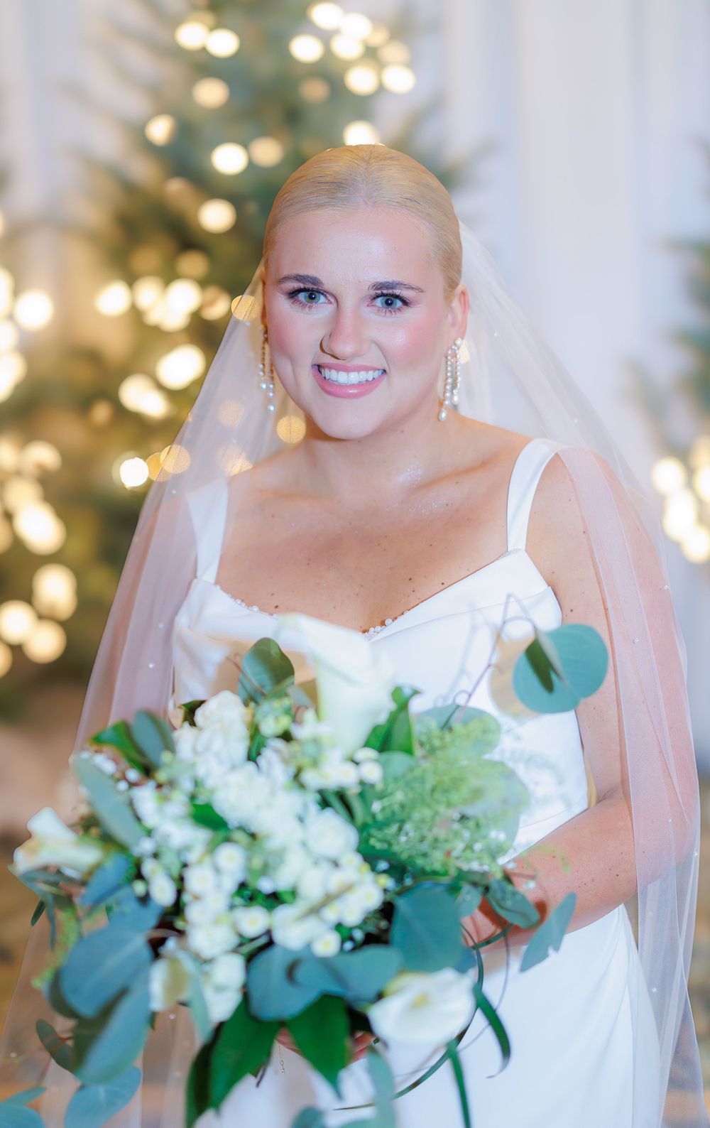 Beautiful bride in white dress holds elegant bouquet with eucalyptus and white flowers against lit Christmas tree.
