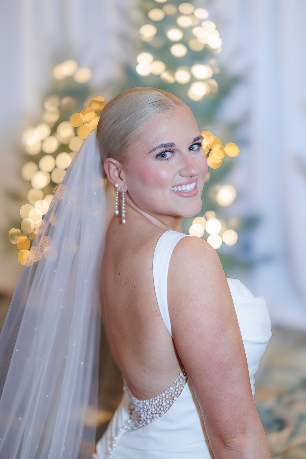 A bride in a white dress with open back and veil poses with Christmas tree lights sparkling in the background.