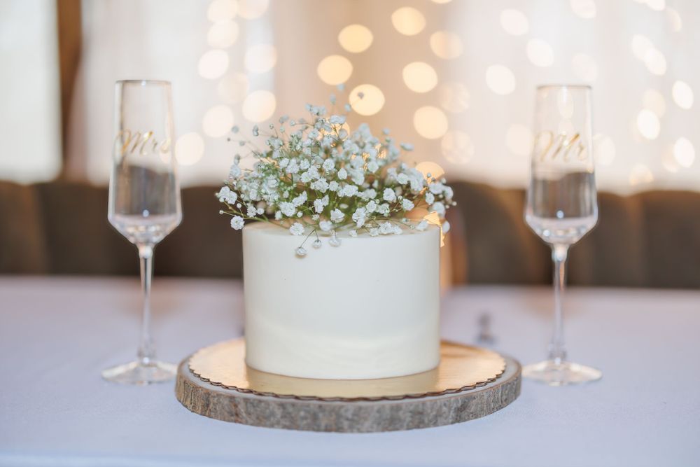 White floral centerpiece on rustic wood slice with two champagne flutes and soft bokeh lights in background.