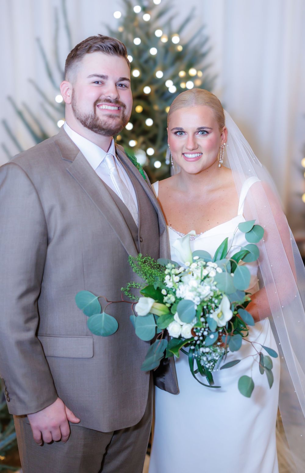 Newlywed couple poses in front of a lit Christmas tree, with bride holding a white and green eucalyptus bouquet.