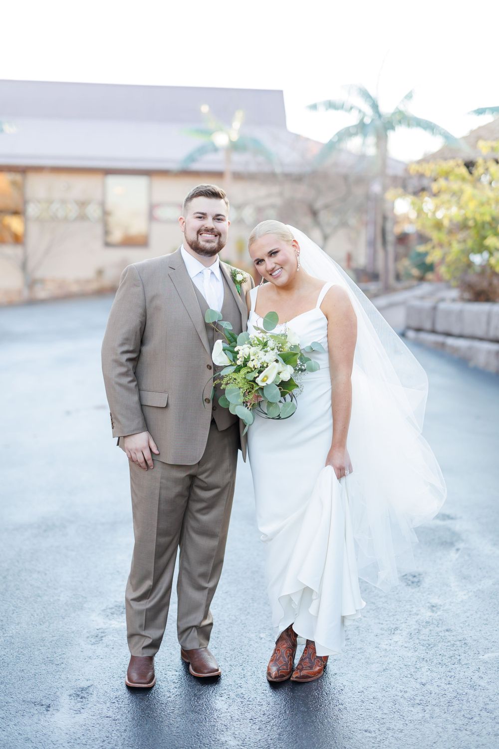 Newly married couple in formal attire poses outdoors on a wet winter day with palm trees in background.