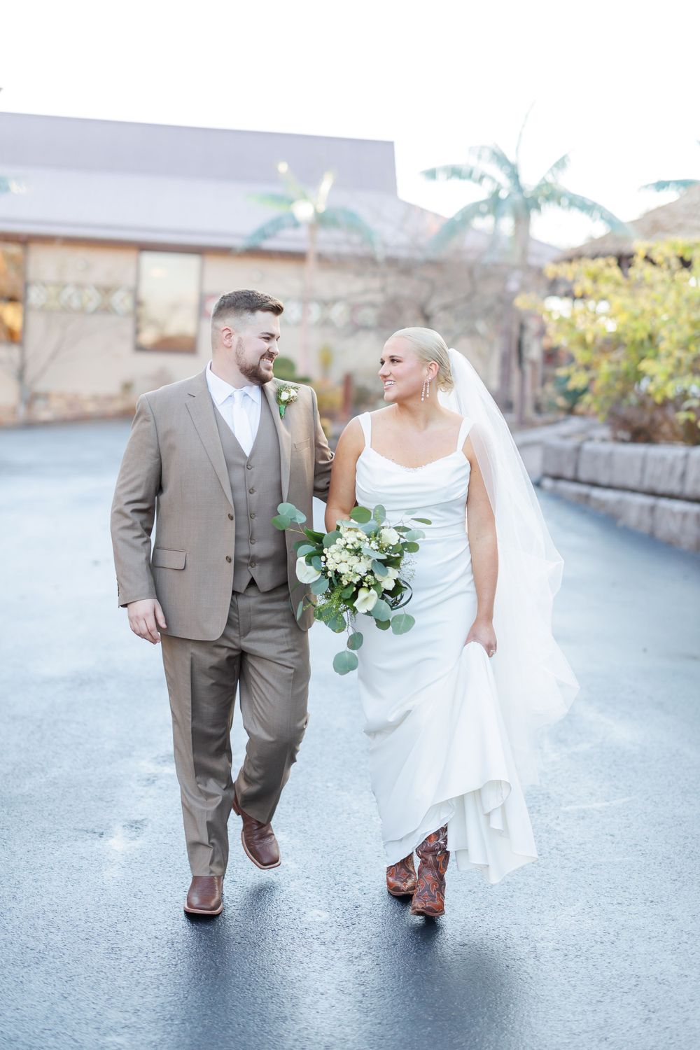 A wedding couple walks and smiles at each other on a paved path with palm trees in the background.
