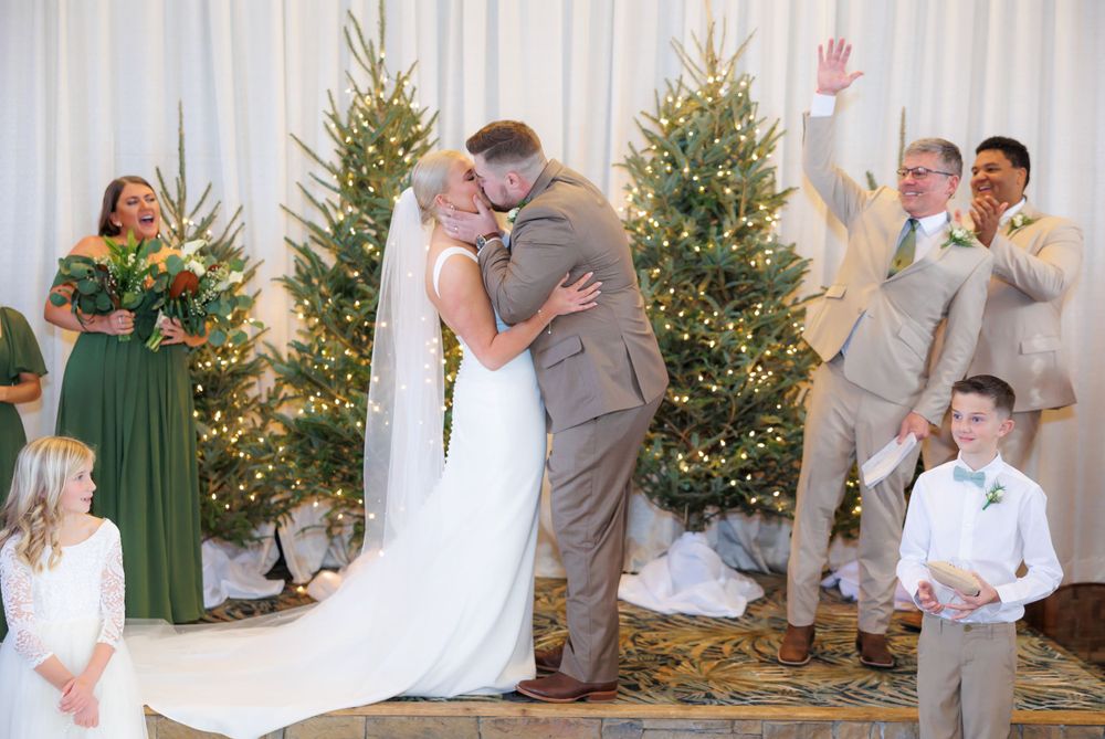 A couple shares their first kiss during a winter wedding ceremony with lit Christmas trees and wedding party looking on.