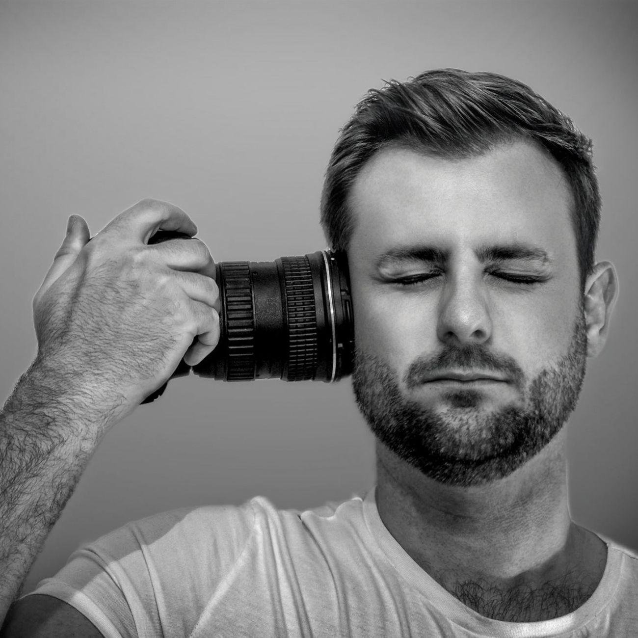 Black and white portrait of a photographer holding a camera lens to face with eyes closed and neutral expression. A headshot.
