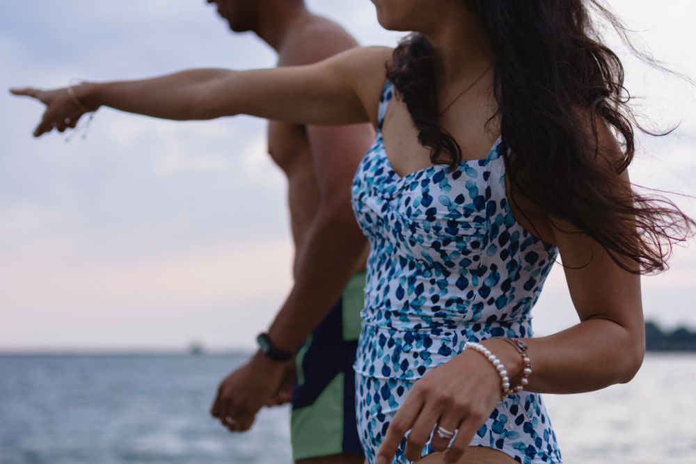 A couple considers jumping into Lake ontario as they stand on a dock in their swimsuits at Lake Ontario. She points ahead at something.