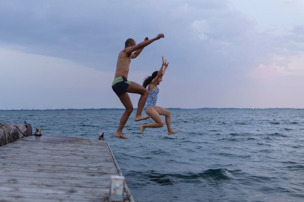 A couple runs/ jumps in the air from a dock into Lake Ontario, in Kingston, Ontario, Canada. Their arms and legs are spread wide in the air.