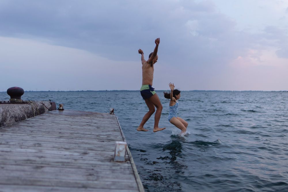 A couple jumps in the air from a dock into Lake Ontario, in Kingston, Ontario, Canada. Their arms and legs are spread wide in the air.